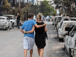 Una pareja camina junto a varios vehículos calcinados en una zona afectada por los incendios en Argyra Akti, en Mati (Grecia). El primer ministro, Alexis Tsipras, declara tres días de luto. EFE / P. Saitas