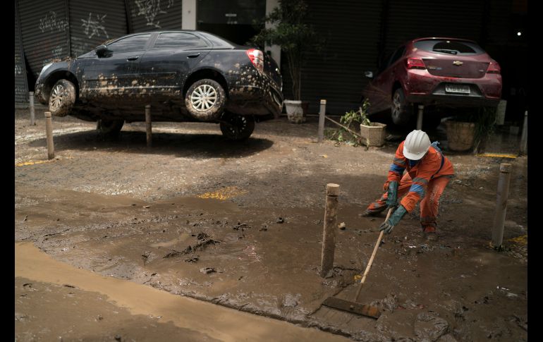 Un trabajador del gobierno quita lodo de una acera junto a carros dañados.
